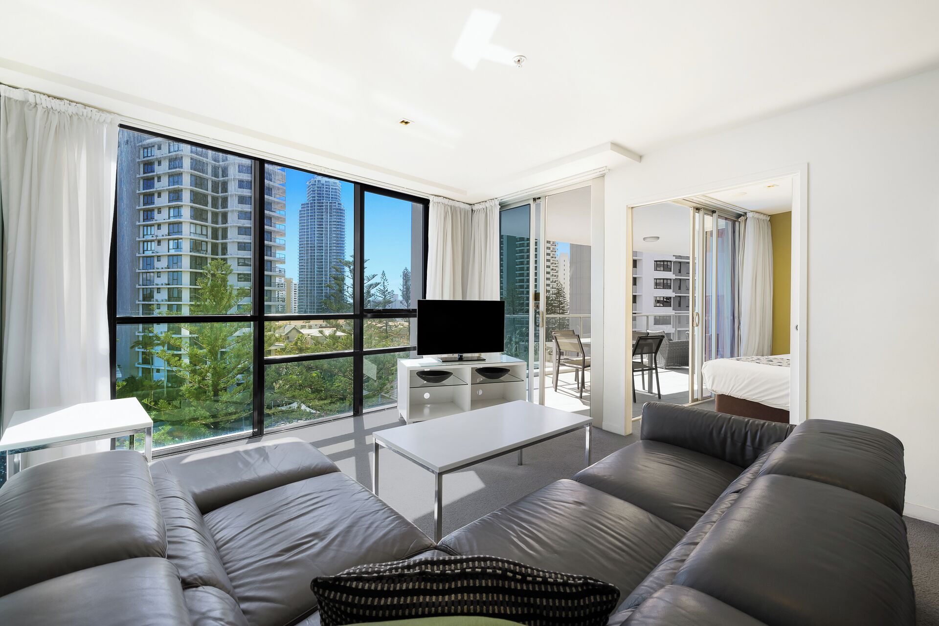 A modern living room with a large window, a flat-screen TV, a sofa, a coffee table, and a balcony with a chair.