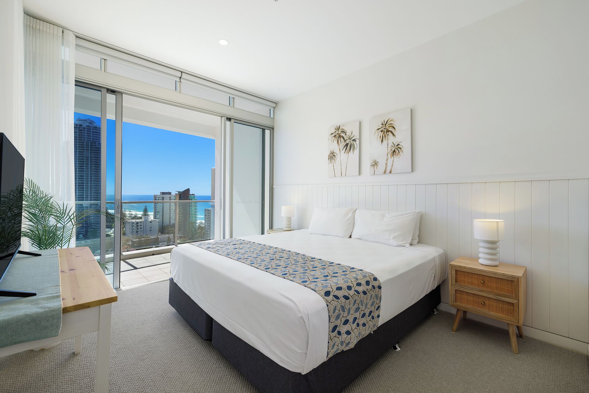 A modern hotel room with a large bed, a desk, a nightstand, and a view of the city and ocean.