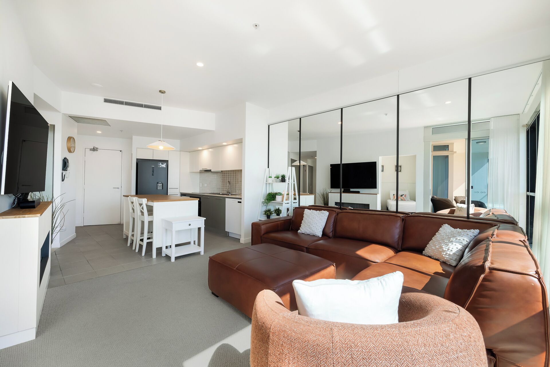 A modern living room with a brown leather sofa, a white dining table with chairs, a kitchen area with white cabinets, and a large mirror reflecting the room.