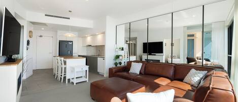 A modern living room with a brown leather sofa, a white dining table with chairs, a kitchen area with white cabinets, and a large mirror reflecting the room.