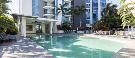 A modern hotel with a swimming pool, sun loungers, and palm trees.