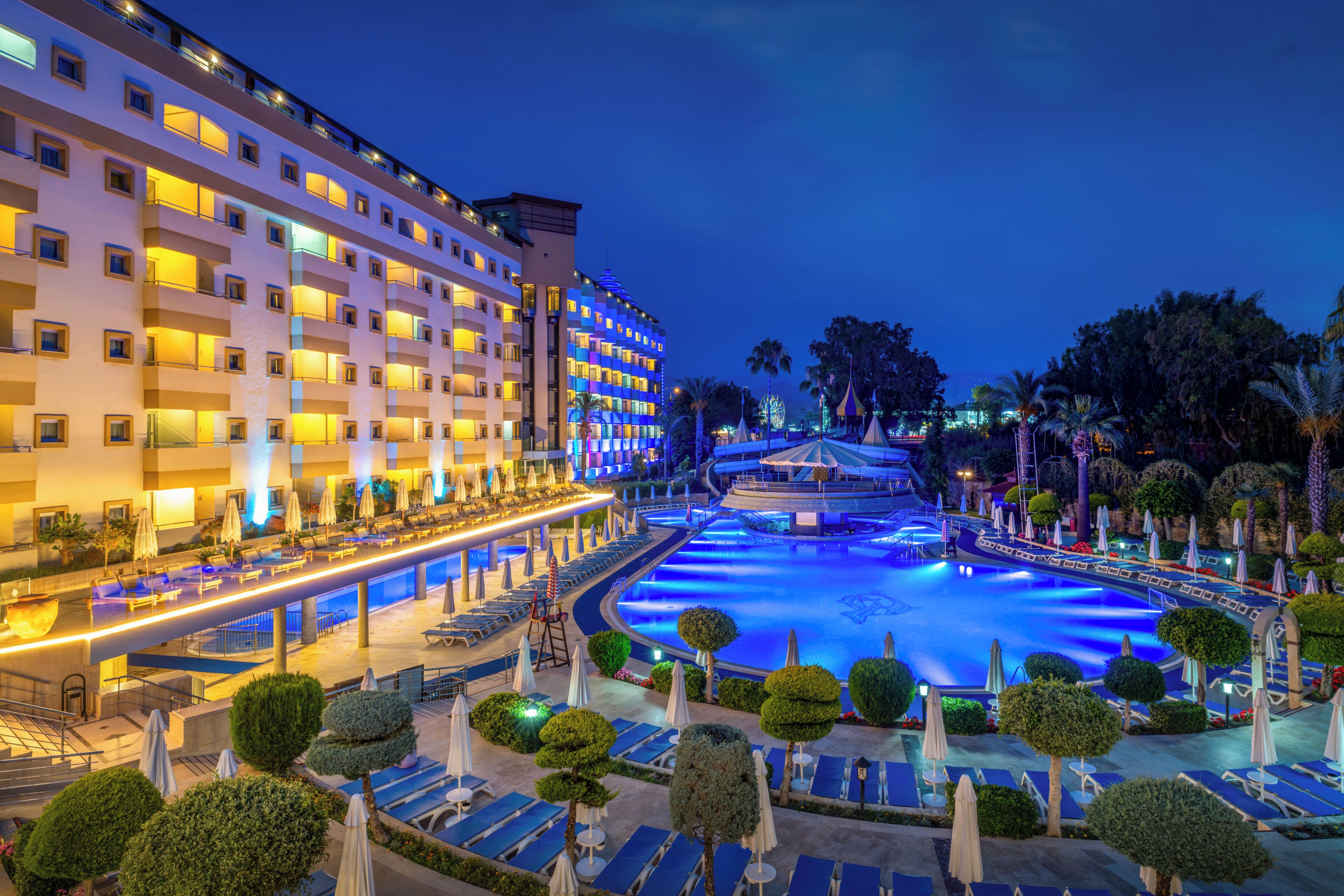 A hotel with multiple floors, a swimming pool, and a well-lit outdoor area with lounge chairs and umbrellas.