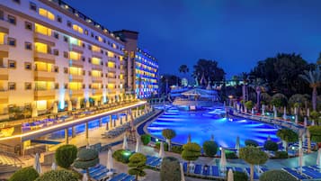 A hotel with multiple floors, a swimming pool, and a well-lit outdoor area with lounge chairs and umbrellas.