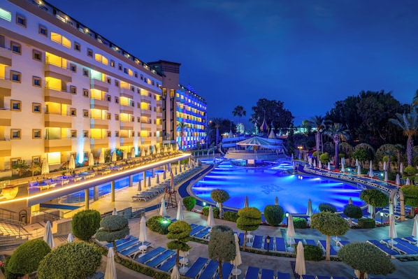 A hotel with multiple floors, a swimming pool, and a well-lit outdoor area with lounge chairs and umbrellas.