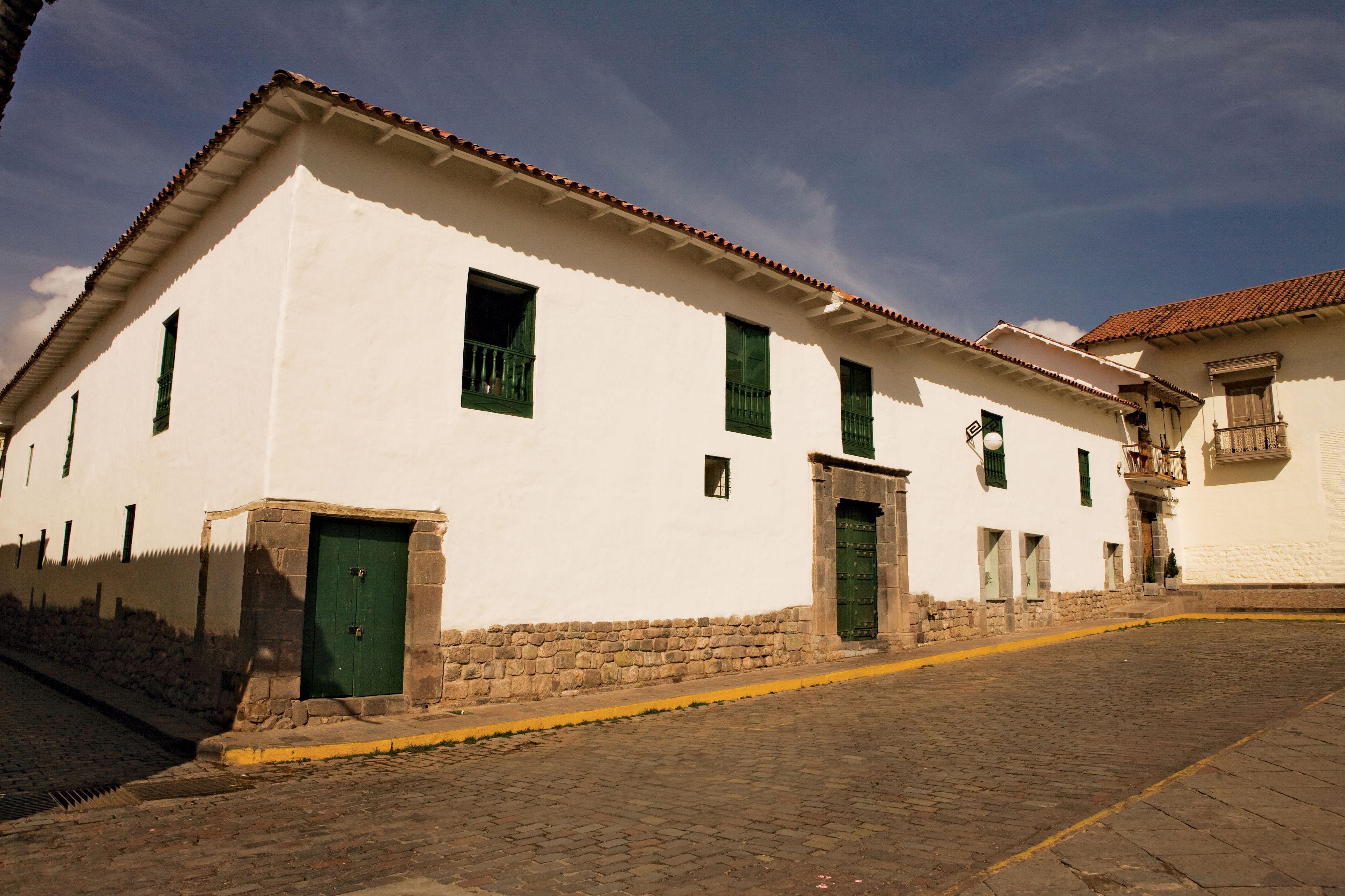 A white building with green doors and windows, a stone base, and a cobblestone street.