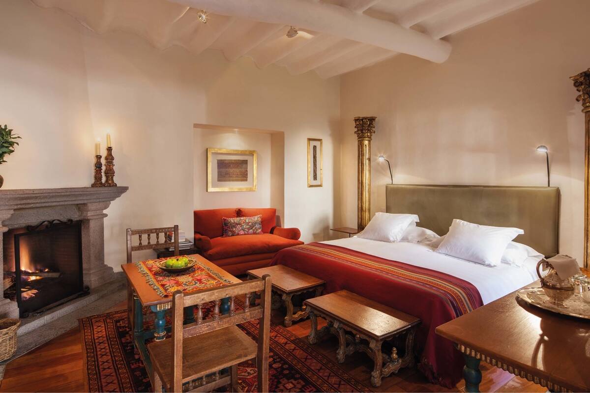 A cozy room with a fireplace, a bed with a headboard, a red sofa, a wooden table, and a patterned rug.