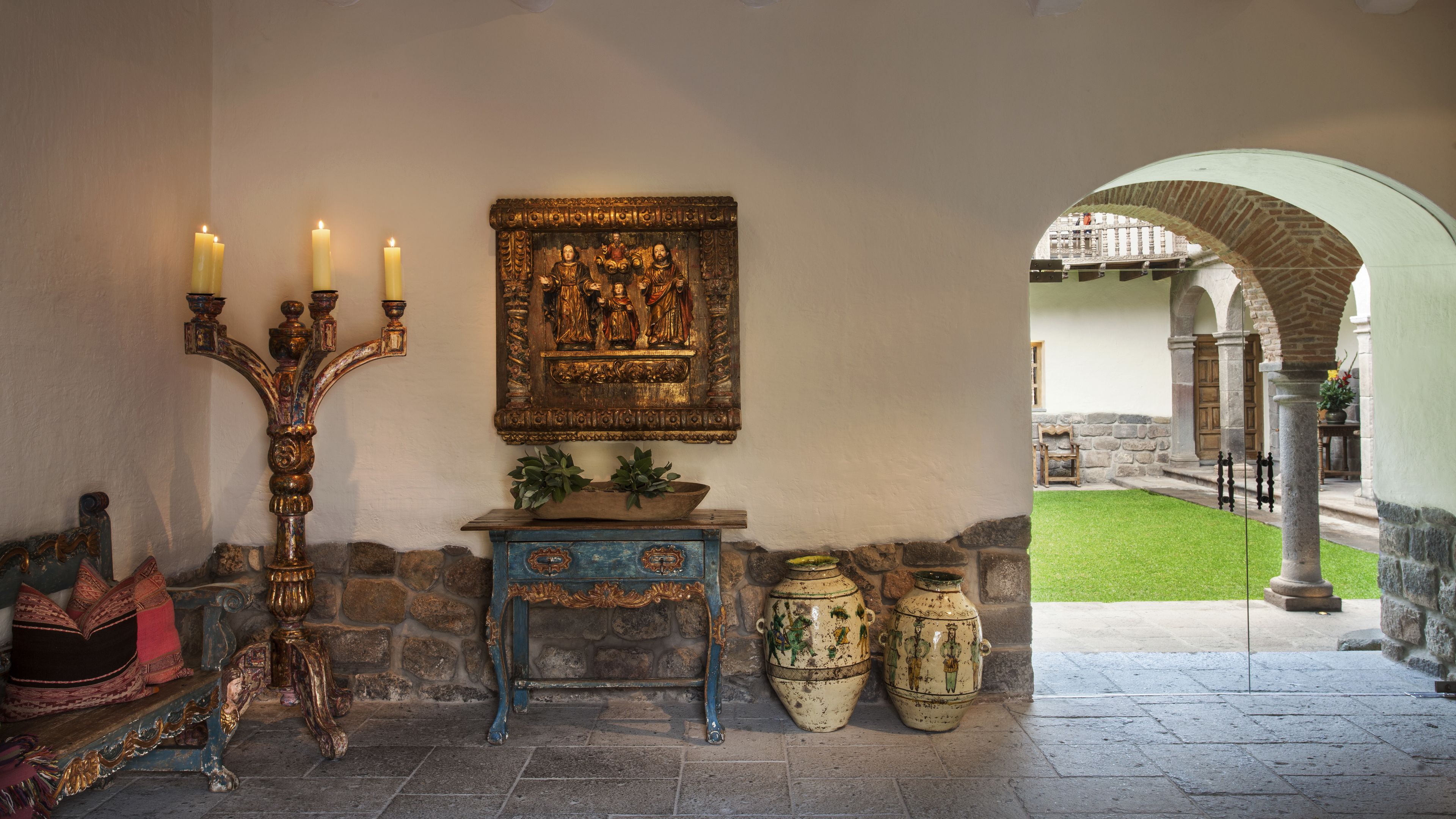 Un muro de piedra con un banco de madera, un candelabro, un cuadro enmarcado y un jarrón grande.