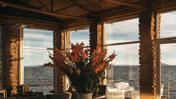 A dining area with a table set for two, a vase of flowers, and a view of the sea through large windows. - Titilaka by Andean (Platería)