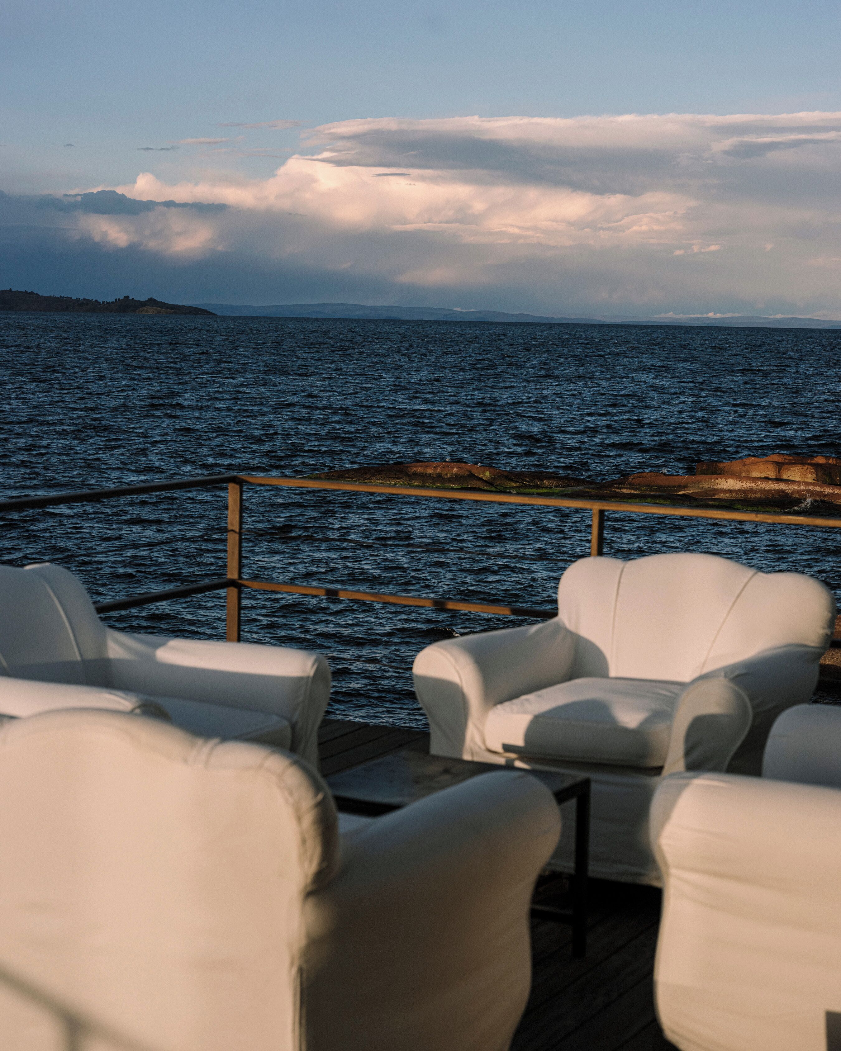 Una terraza con sillas blancas que da a una masa de agua, con una costa rocosa en la distancia.