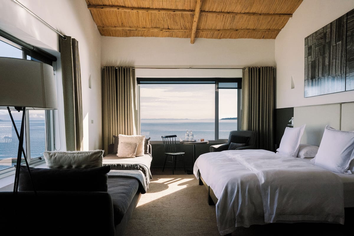 Habitación de hotel con dos camas, un escritorio, una silla y un amplio ventanal con vistas al mar.