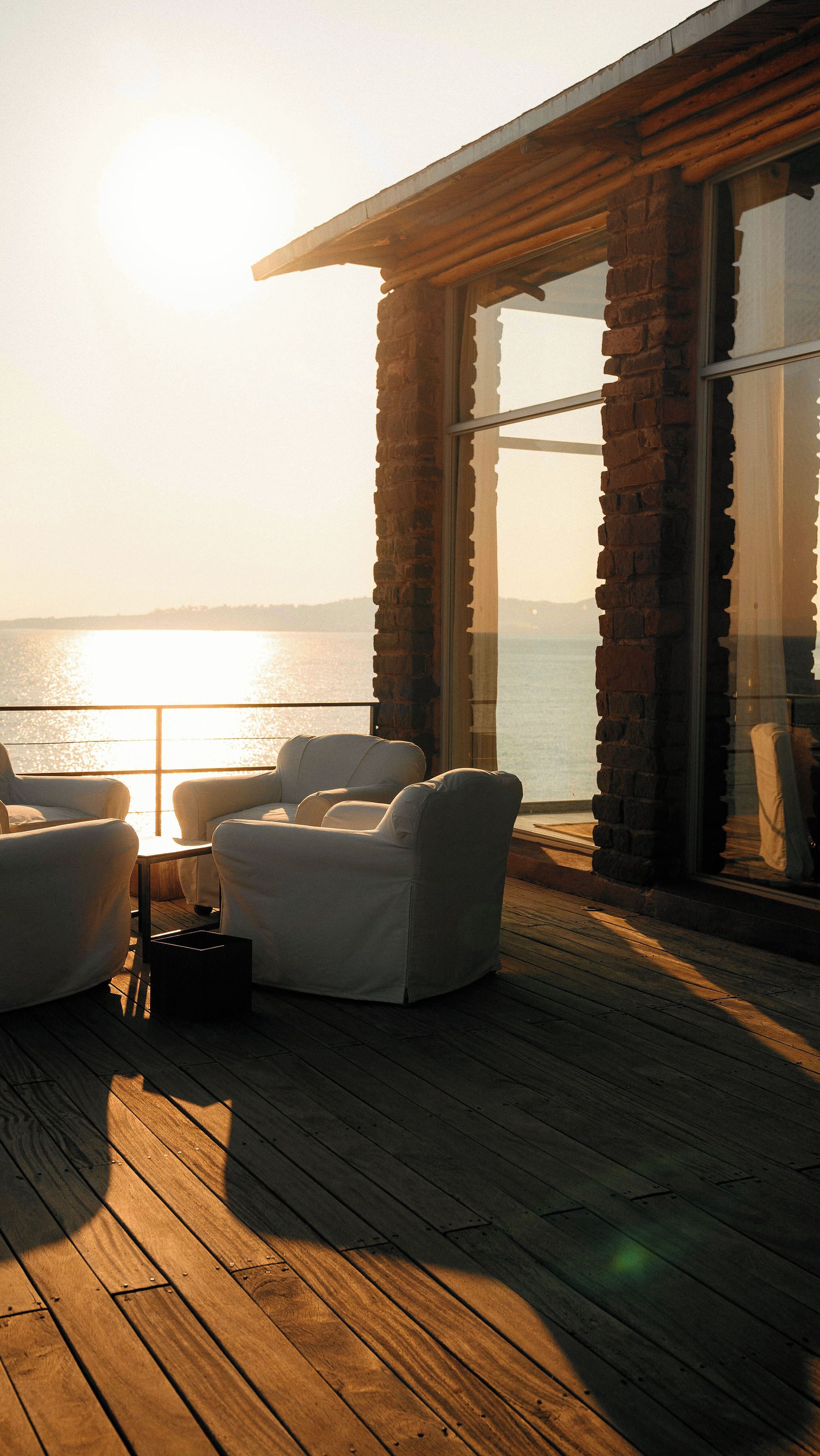 Terraza de madera con dos sillones blancos y una mesa, con vista a un cuerpo de agua y horizonte despejado.