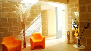 A room with a brick wall, two orange chairs, a tall vase with dried plants, a mirror, and a sculpture. - Cave Bianche Hotel (Favignana)
