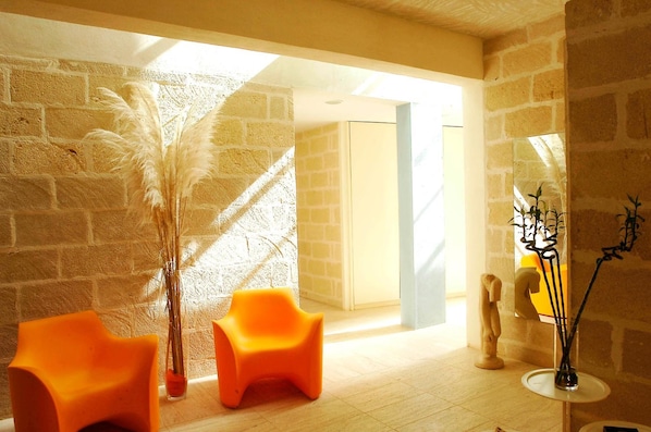 Una habitación con pared de ladrillo, dos sillas naranjas, un jarrón alto con plantas secas, un espejo y una escultura.