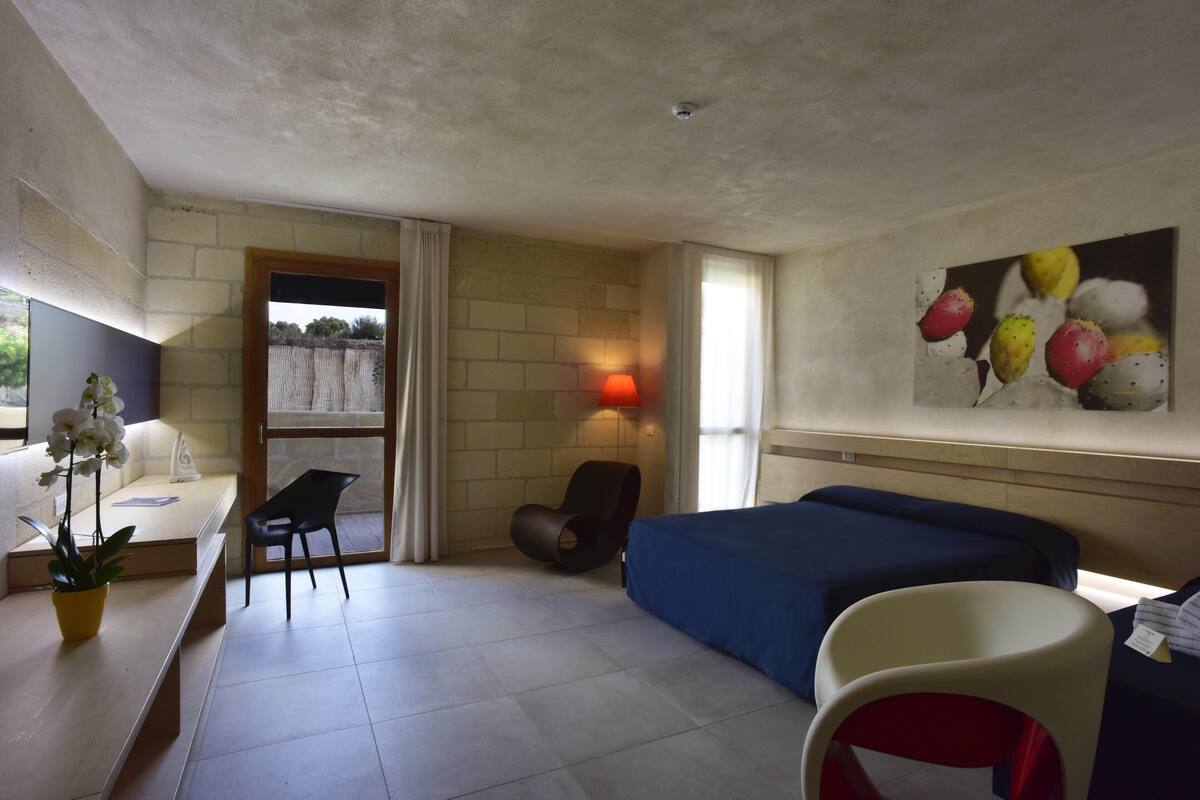 Une chambre d’hôtel avec un lit, un bureau, une chaise, une plante en pot et un tableau au mur.