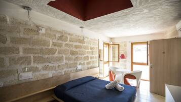 Une pièce avec un mur en briques, un lit recouvert d’une couverture bleue, un banc en bois, une table blanche avec une chaise rouge et une porte en bois.