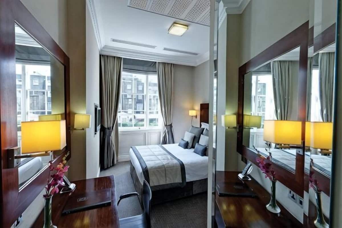 A hotel room with a bed, bedside tables, lamps, a mirror, a window with curtains, and a view of buildings.