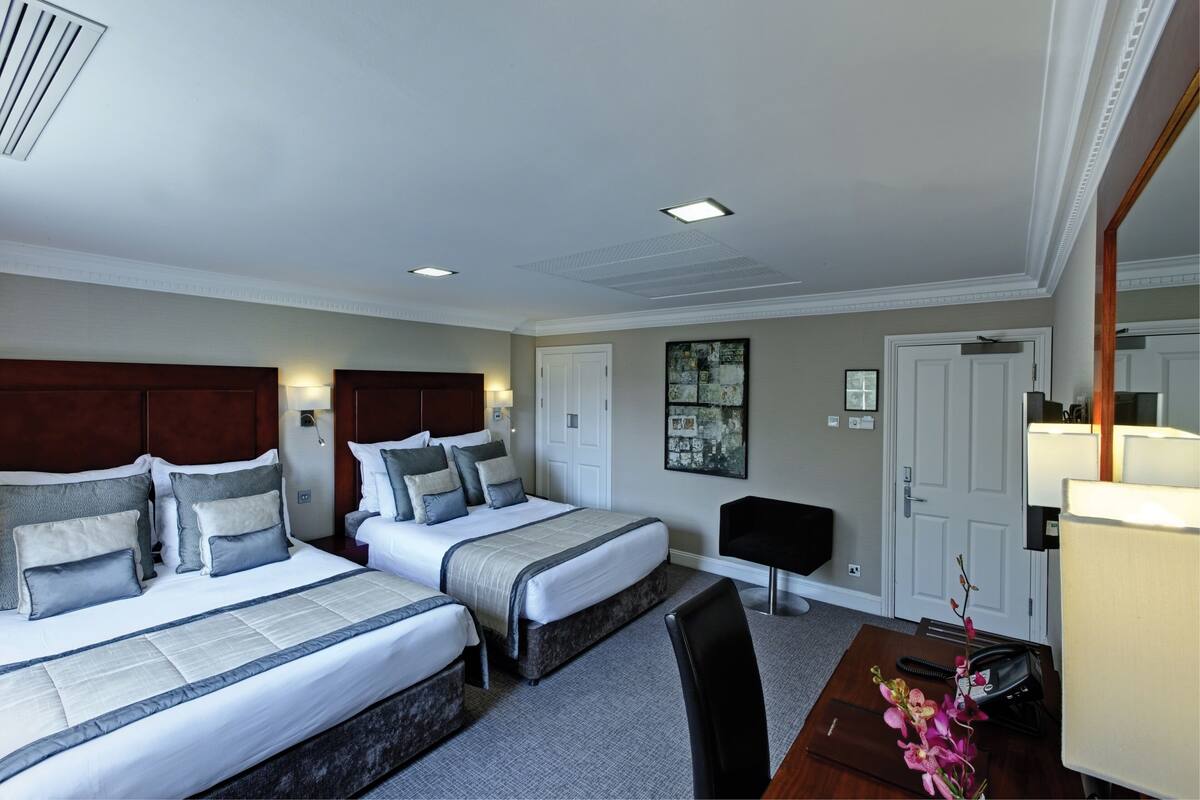 A hotel room with two beds, a desk, a chair, a lamp, and a door.