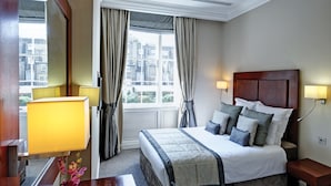 A hotel room with a large bed, two bedside lamps, a window with curtains, and a view of buildings outside. - Grange Beauchamp (London)
