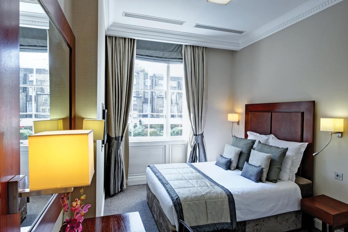 A hotel room with a large bed, two bedside lamps, a window with curtains, and a view of buildings outside.
