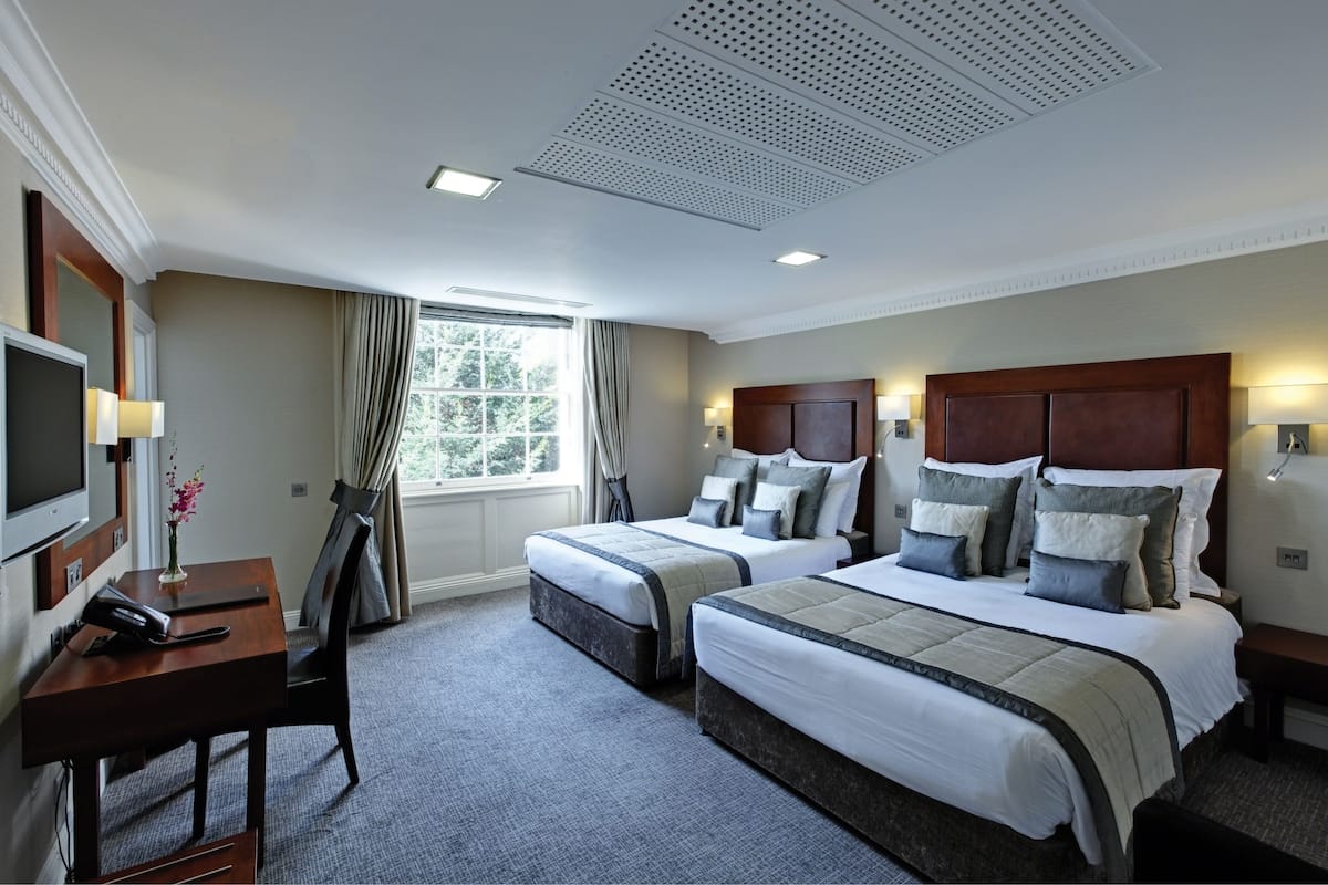 A hotel room with two beds, a desk, a TV, and a window with curtains.