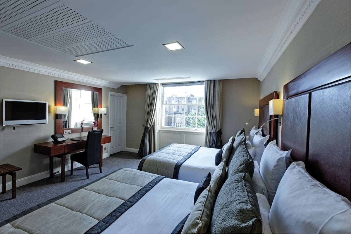 A hotel room with two beds, a desk, a chair, a TV, and a large window with curtains.