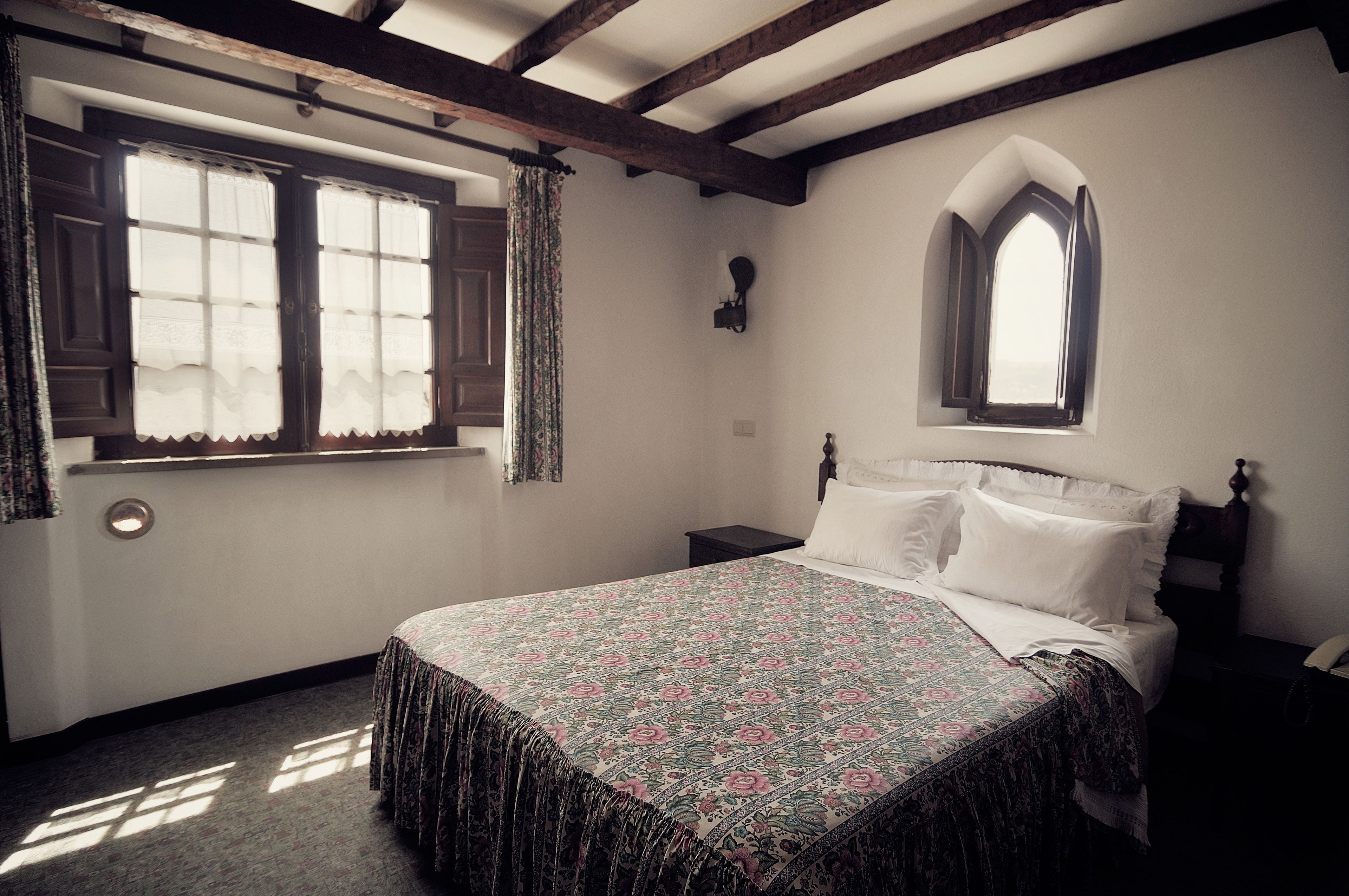 A bedroom with a bed, a window with curtains, a wooden headboard, and a small wall-mounted light.