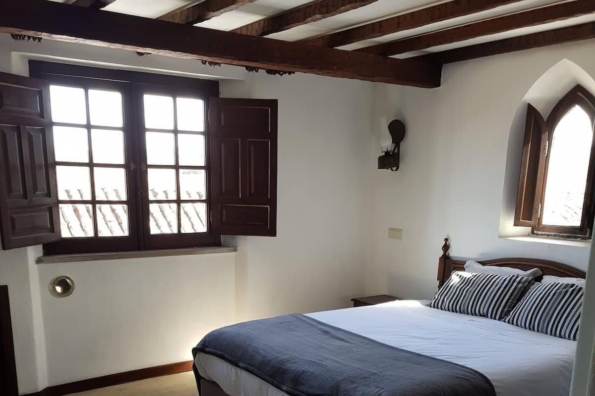 Une chambre avec un grand lit, des poutres en bois au plafond, une fenêtre avec des volets et une petite table placée à côté du lit.