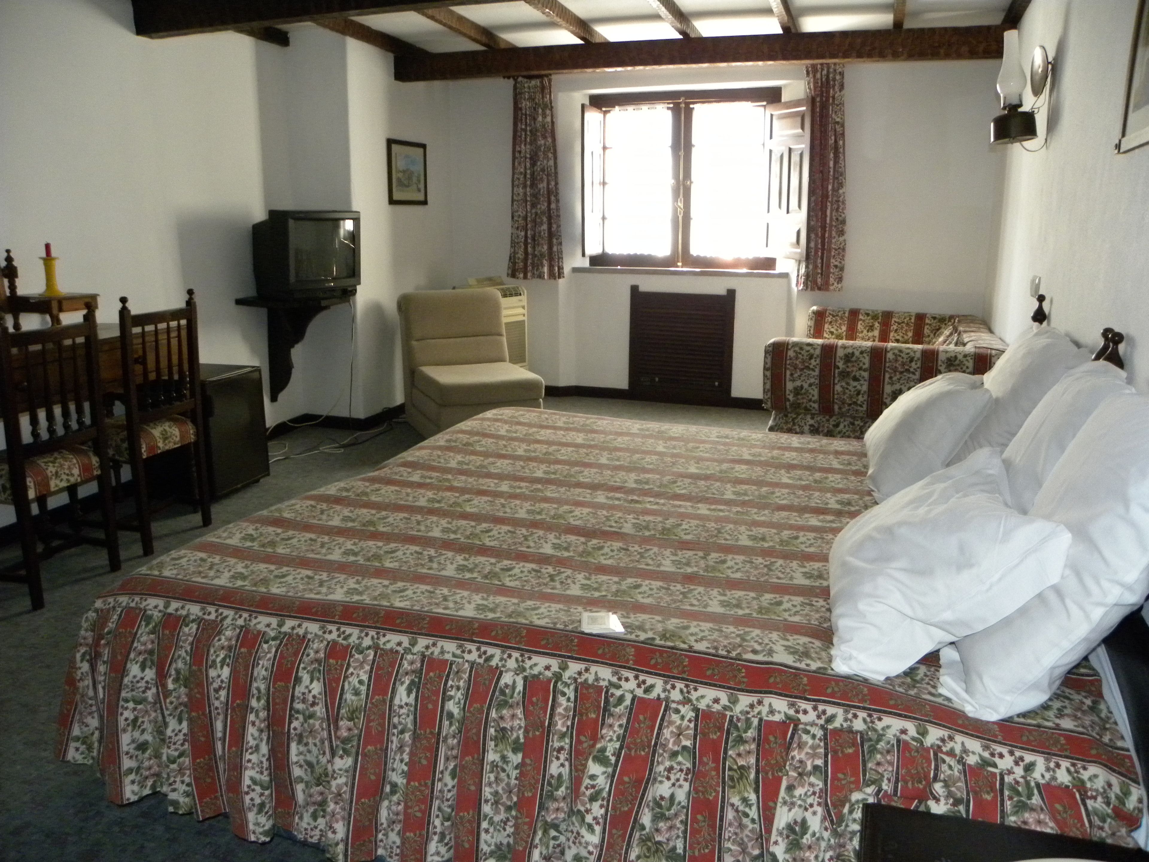 A bedroom with a large bed, a chair, a small table, a television, and a window with curtains.
