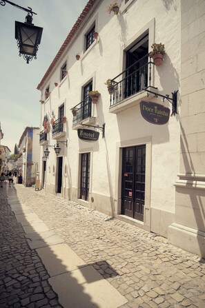 A cobblestone street with a hotel named "Hotel Doce Rainha" and a building with balconies adorned with potted plants. - Hotel Rainha Santa Isabel (Obidos)