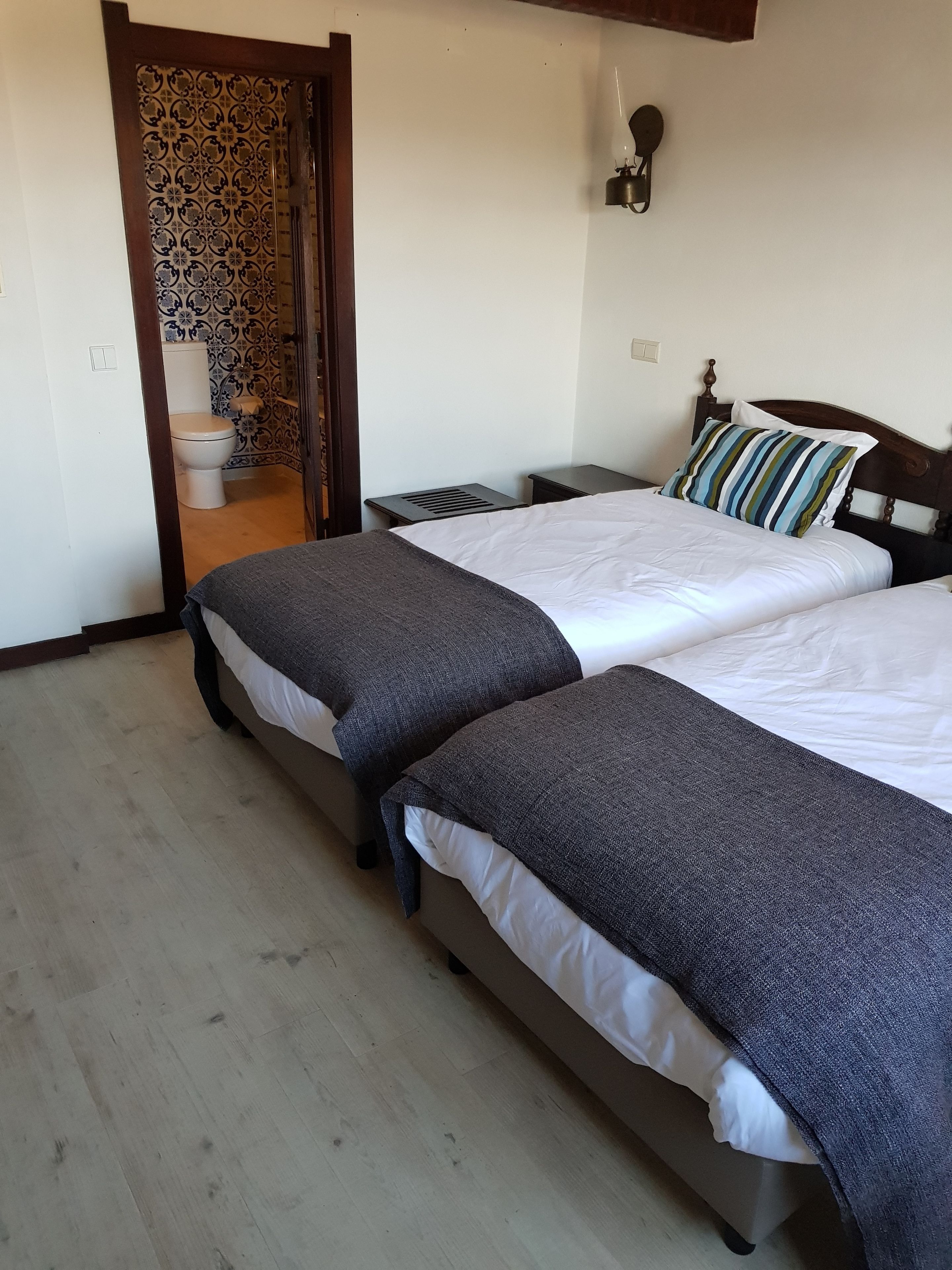 A hotel room with two beds, a wooden floor, a white wall, a wooden door, and a bathroom visible through an open door.