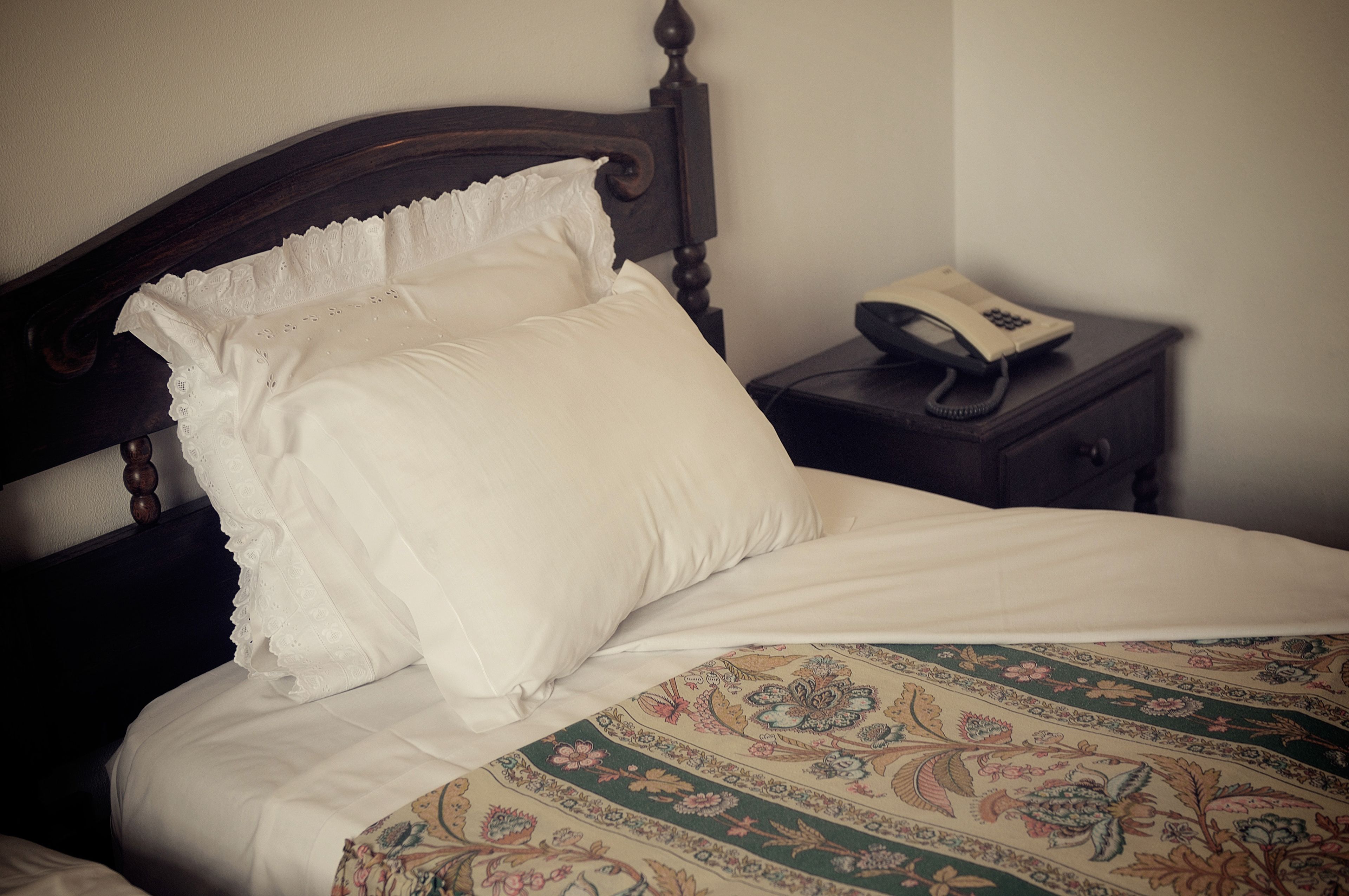 A bed with white linens and a patterned bedspread, a wooden headboard, and a bedside table with a telephone.