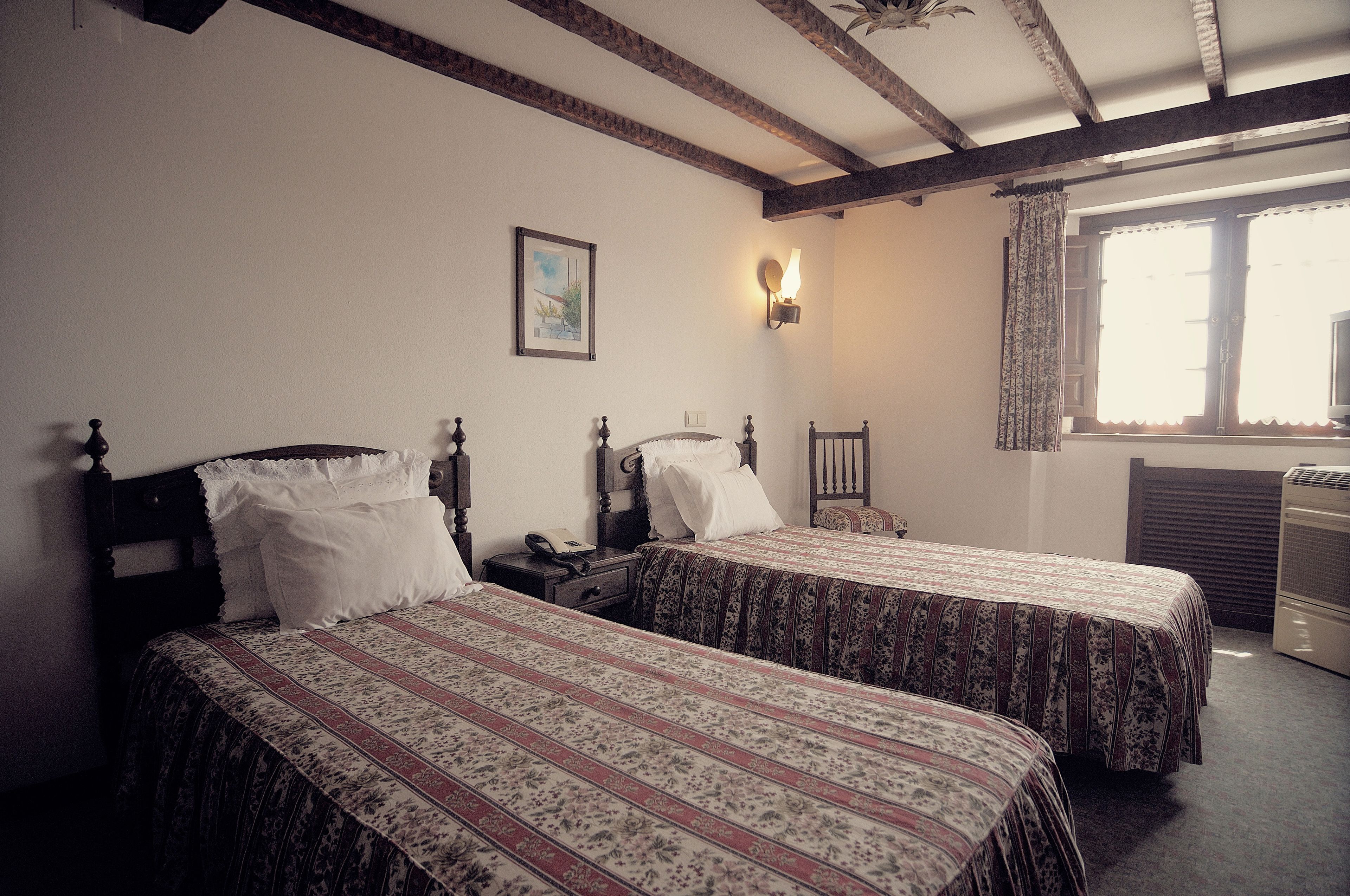 A room with two beds, a nightstand, a lamp, a chair, and a window with curtains.