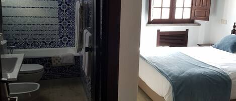 Una habitación con piso de madera, una cama con manta azul, una ventana con contraventanas de madera, y un baño con azulejos estampados y un inodoro.