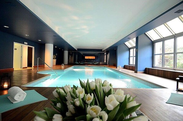 A large indoor swimming pool with a clear view of the sky through skylights, surrounded by wooden flooring and a blue wall.