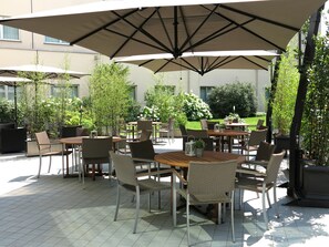An outdoor patio with multiple tables and chairs, shaded by large umbrellas, surrounded by greenery and a building in the background.