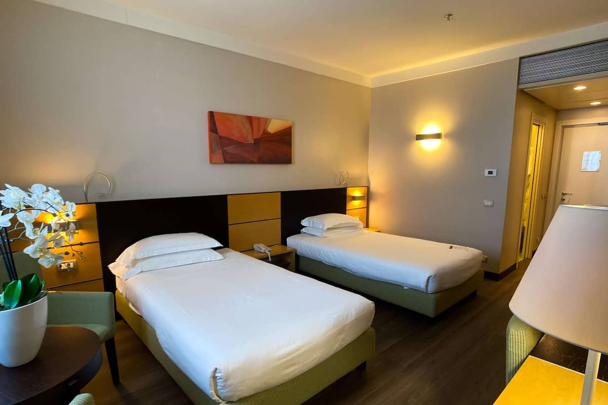 Ett hotellrum med två sängar, ett skrivbord, en stol, en lampa, en krukväxt och en tavla på väggen.