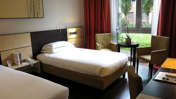 A hotel room with a bed, bedside tables, a desk, chairs, a window with a view of greenery, and a wall-mounted artwork.
