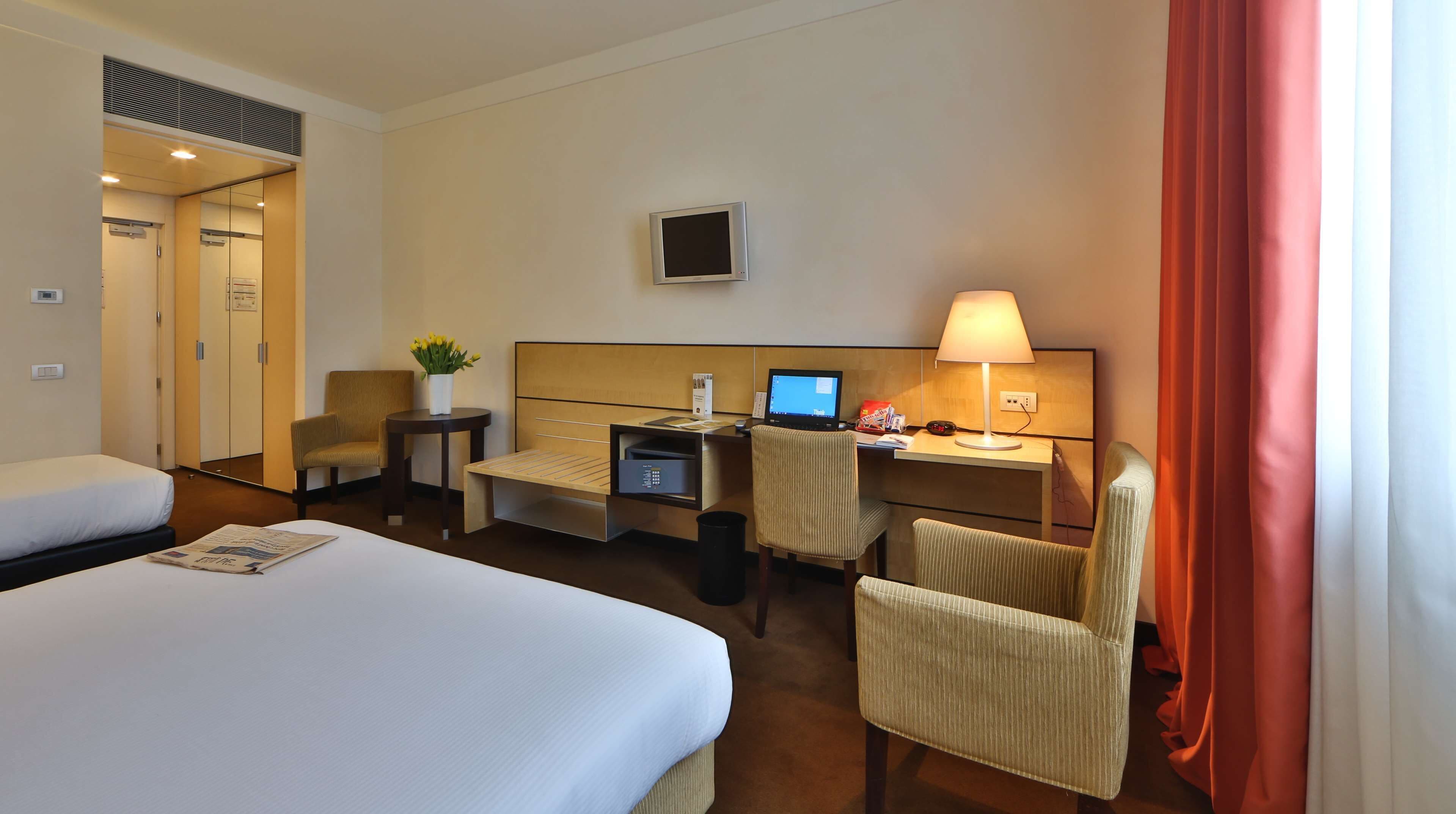 Habitación de hotel con una cama grande, un escritorio con computadora, una silla, una mesita con flores y un televisor en la pared.