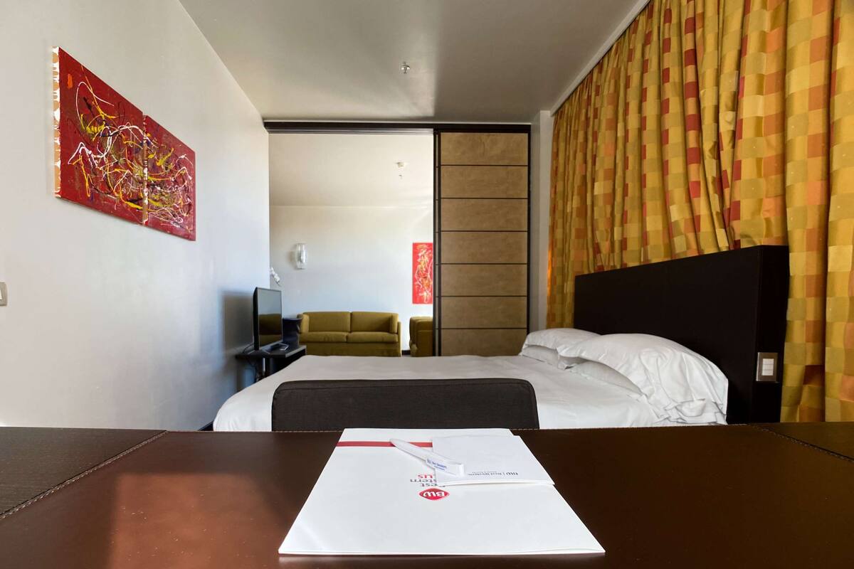 Ett hotellrum med en säng, ett skrivbord med en dator, en tv, en soffa och en tavla på väggen.