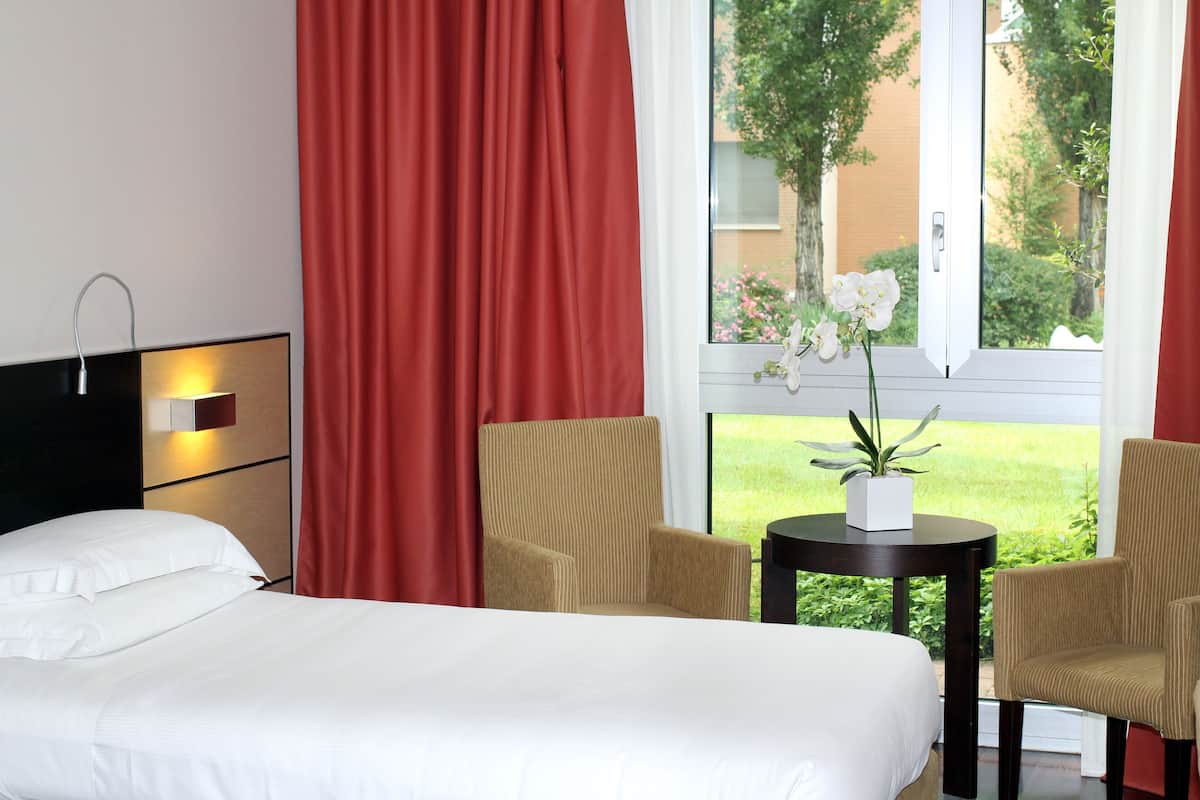 Ett hotellrum med en säng, två stolar, ett litet bord med en växt och ett fönster med utsikt över grönområden.