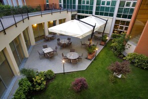 An outdoor patio area with tables and chairs, surrounded by greenery and a large umbrella.