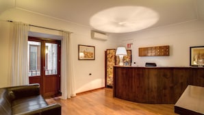 A room with a wooden floor, a leather sofa, a wooden reception desk, and a window with curtains. - Residenza Domizia- Guest House (Rome)