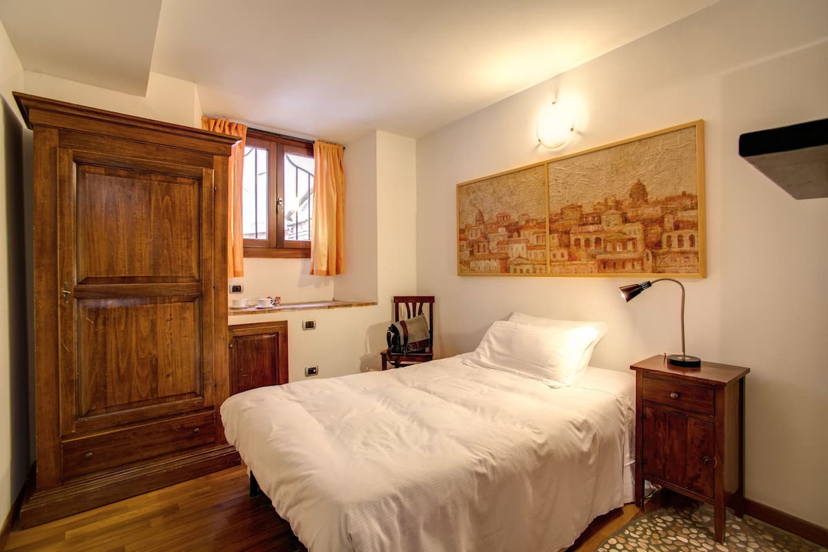 A bedroom with a wooden wardrobe, a bed with white bedding, a nightstand with a lamp, a chair, and a painting on the wall.