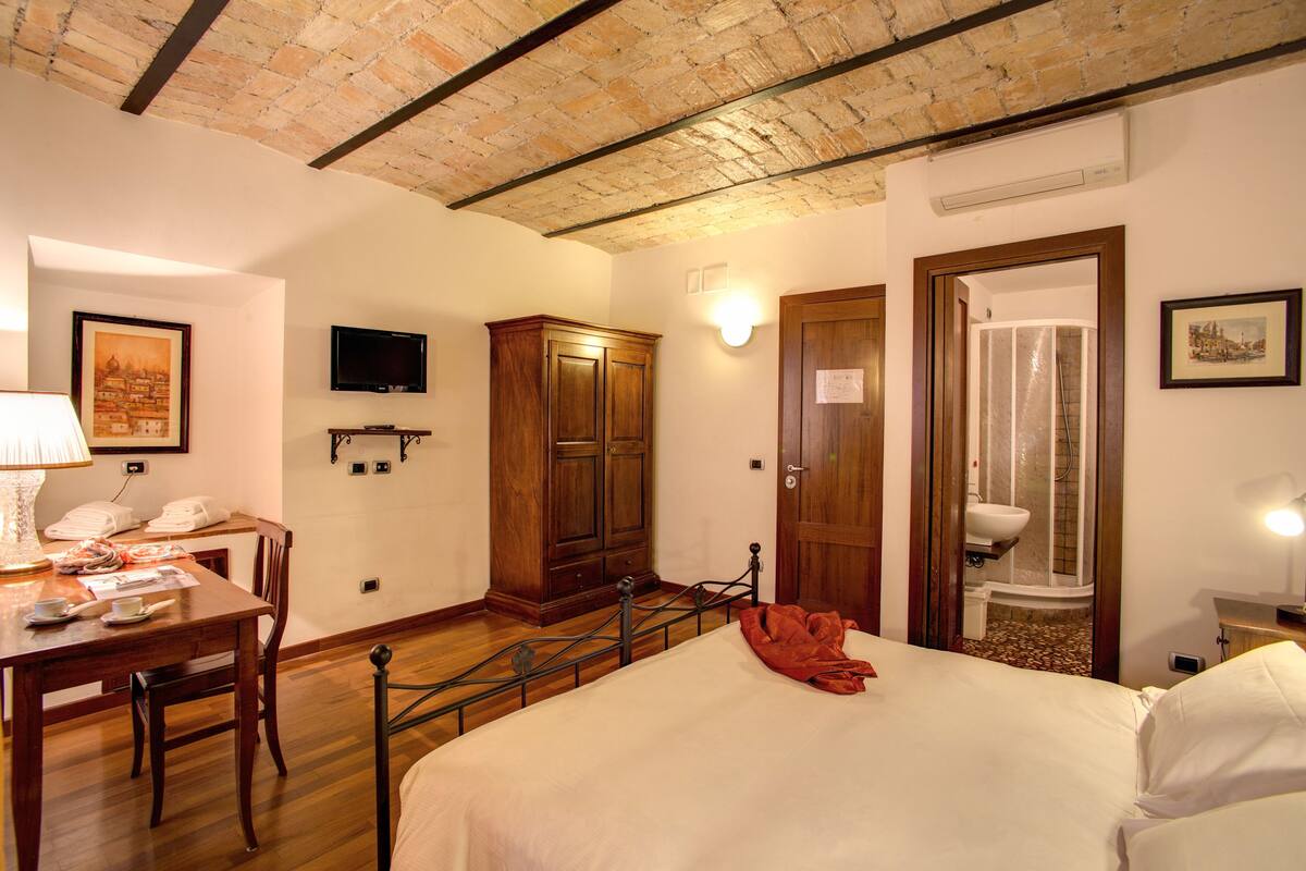 A hotel room with a bed, a wooden wardrobe, a desk with a lamp, a television, and a bathroom visible through an open door.
