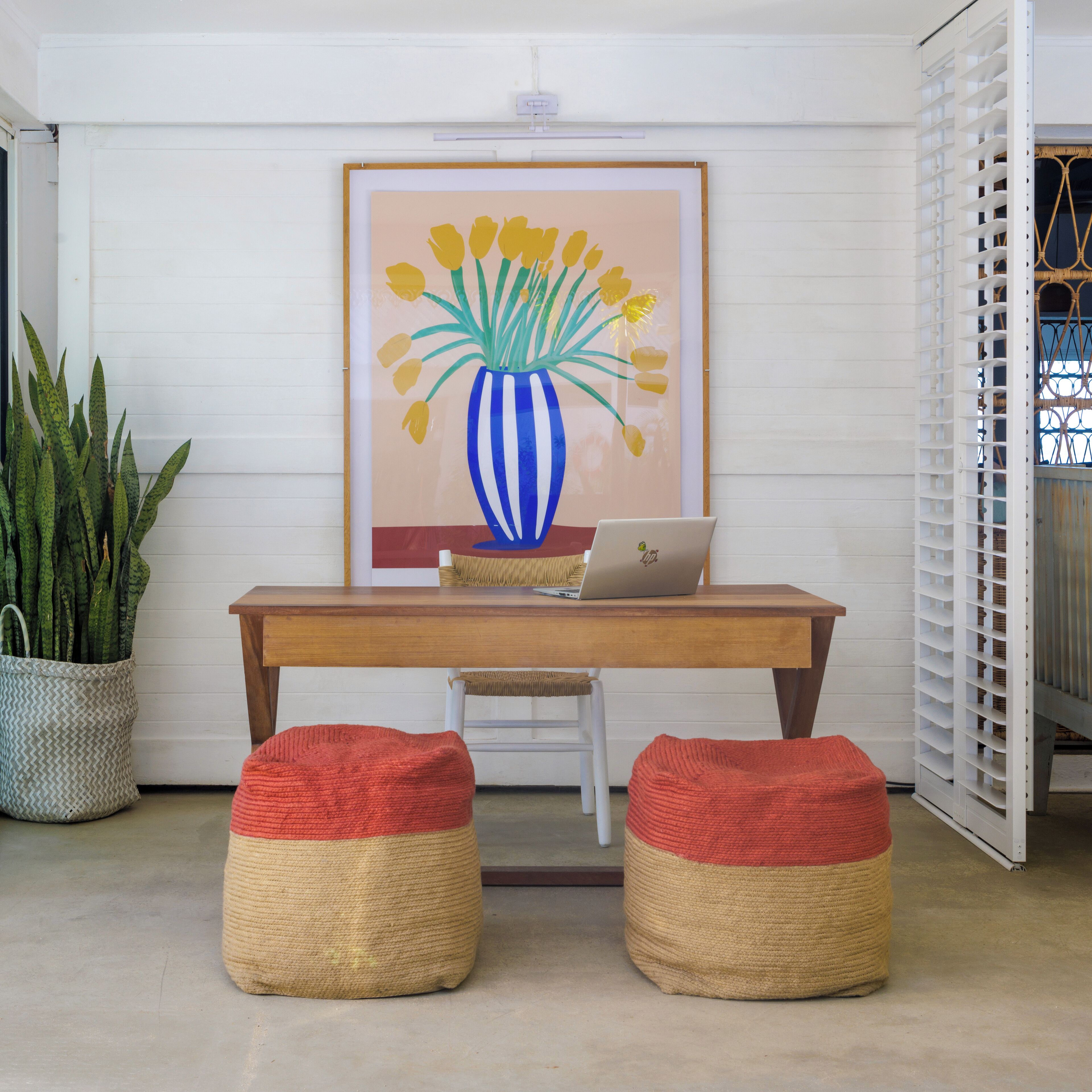 A room with a wooden desk, a laptop, two woven poufs, a potted plant, and a framed artwork on the wall.