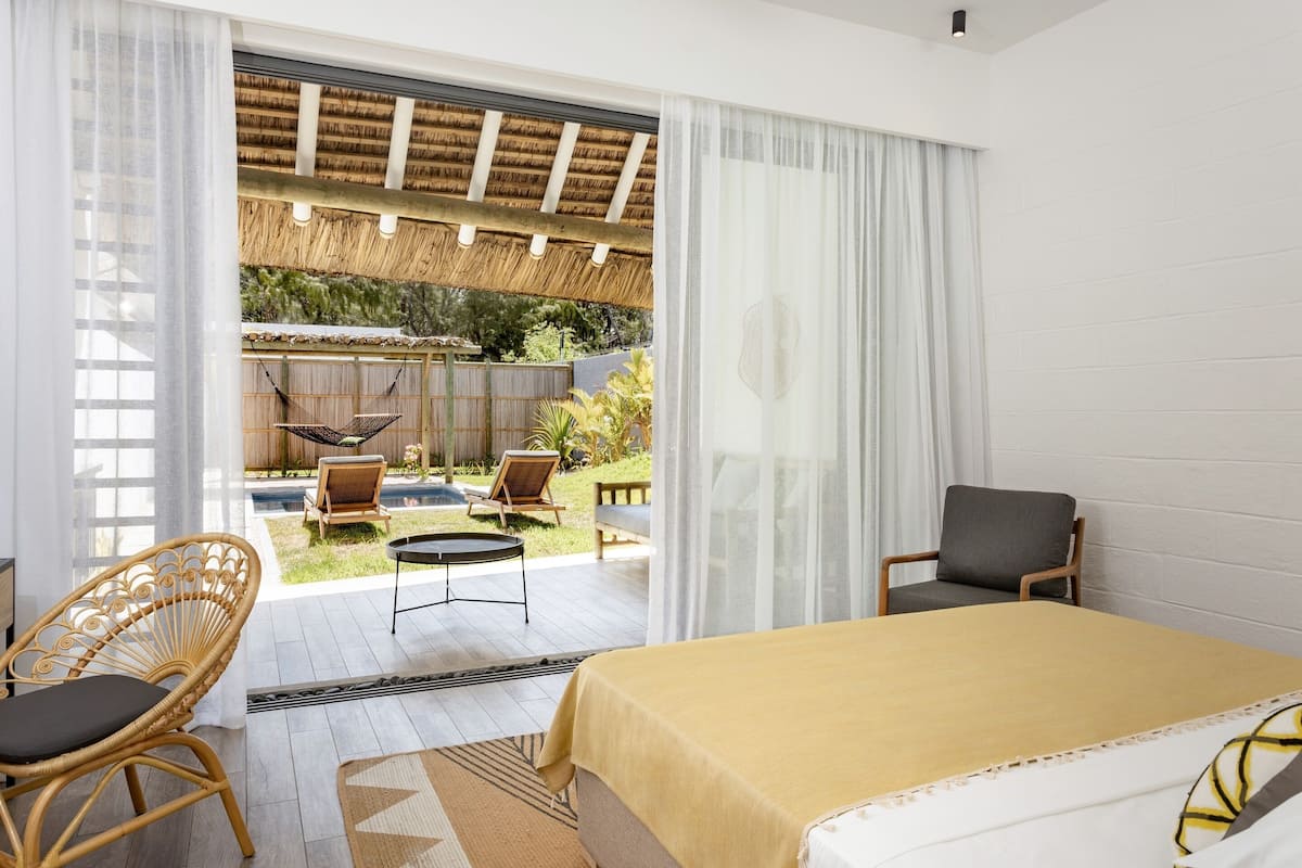 A bedroom with a bed, a chair, a small table, and a view of an outdoor area with a pool and garden.