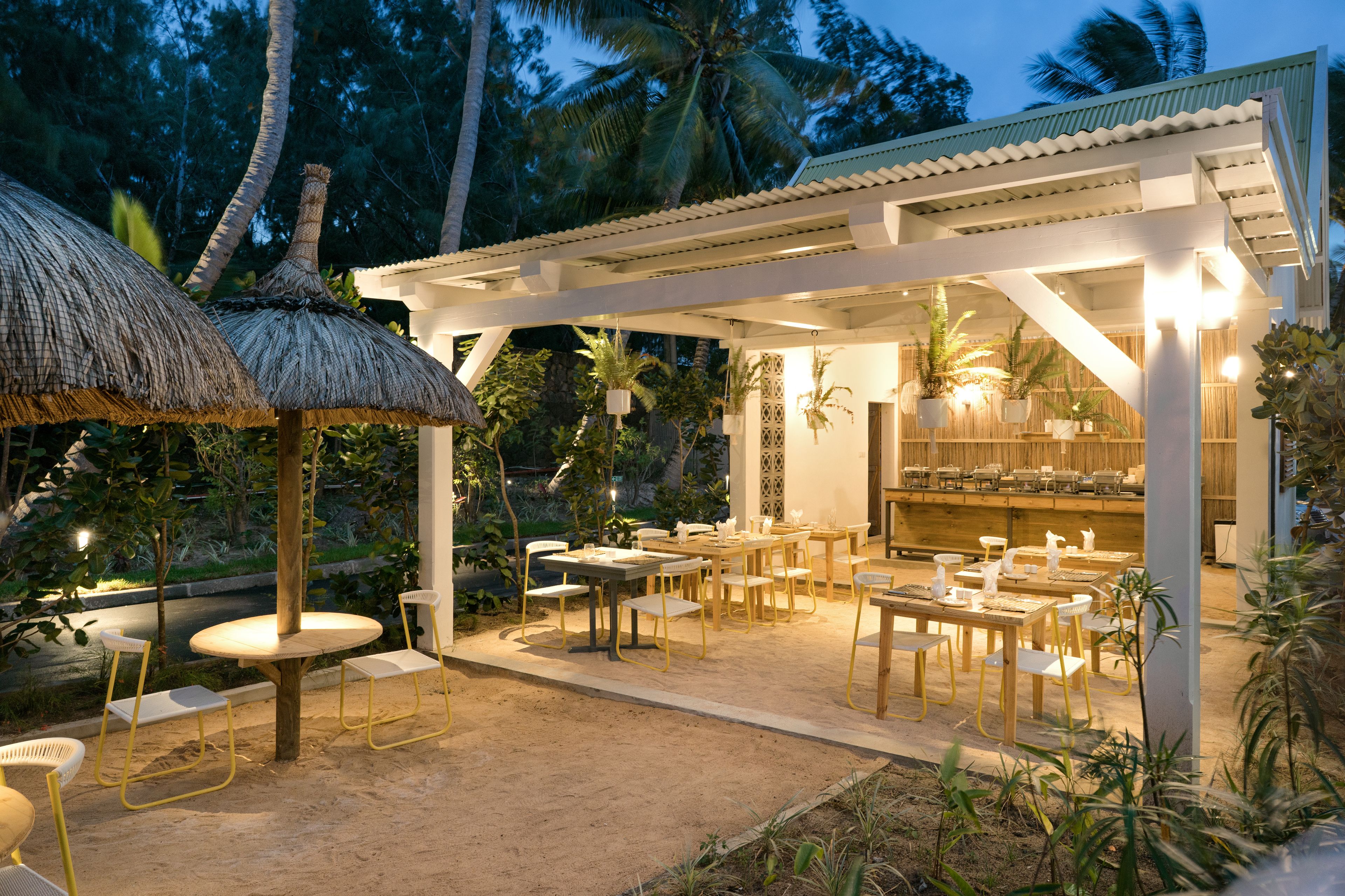 An outdoor dining area with round tables and chairs, surrounded by plants and a thatched-roof structure.
