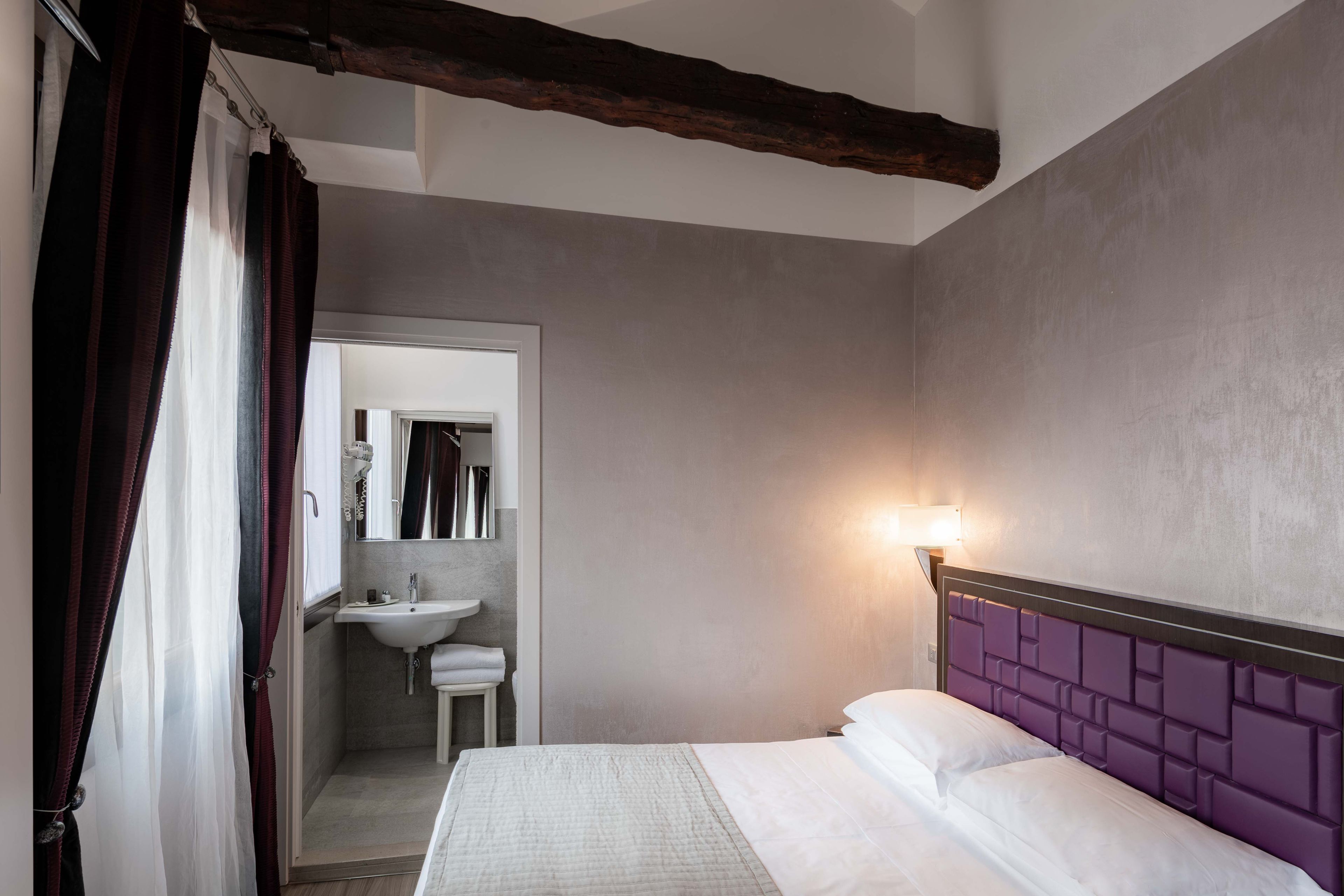 A bedroom with a bed, a nightstand, a bedside table, a window with curtains, and a bathroom visible through an open door.