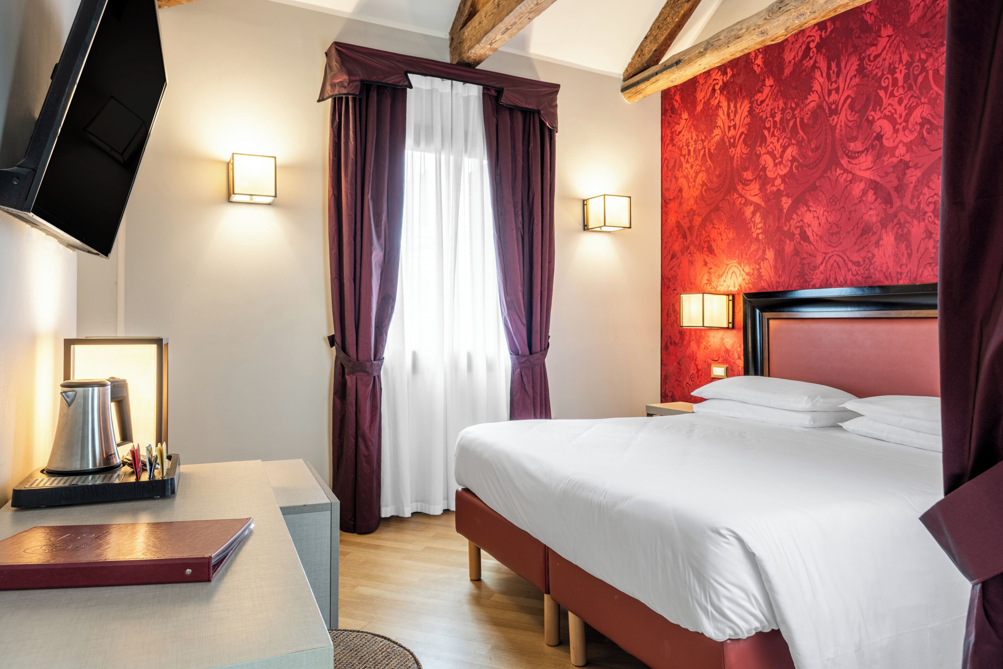 A hotel room with a bed, a desk with a kettle, a flat-screen TV mounted on the wall, and a window with curtains.