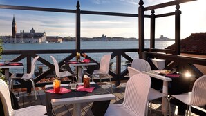 A terrace with tables and chairs overlooking a body of water with historic buildings in the background. - Hotel Paganelli (Venice)